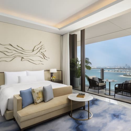 Luxury hotel room with a balcony overlooking a city skyline and water, featuring a bed, side tables with lamps, a sofa with cushions, and modern decor.