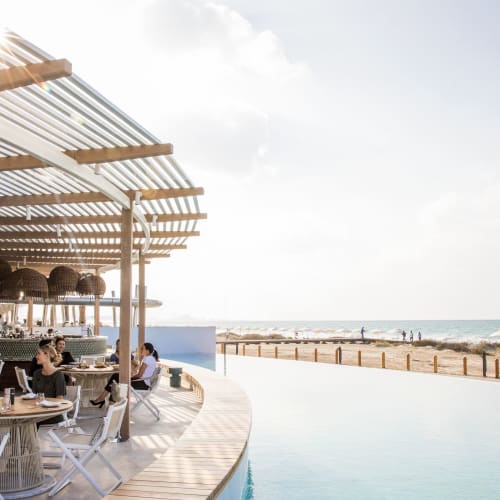 Guests dine at an outdoor poolside restaurant on a sunny beachside resort with ocean views.