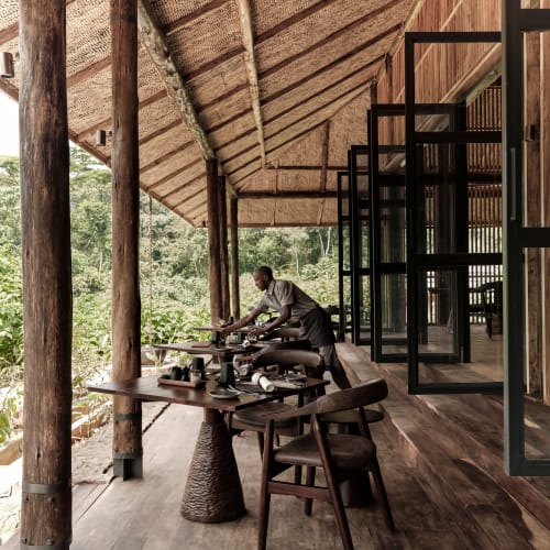 A waiter setting the table on a wooden outdoor terrace at a jungle lodge, with open windows and lush greenery in the background.