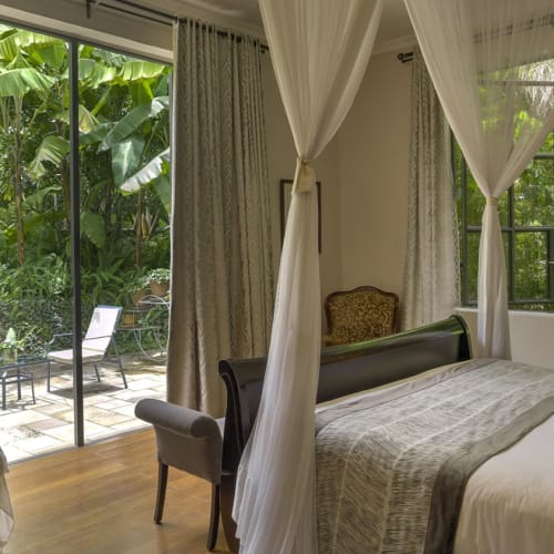 Ein Superior-Gästezimmer mit Himmelbett und weißen Vorhängen, einem Sessel und Schiebetüren aus Glas, die auf eine Terrasse mit üppigem Grün öffnen.