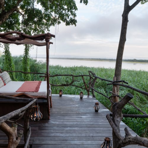 Ein gemütliches Sternbett auf einer Holzbalkon mit Blick auf einen Fluss und grüne Natur in Uganda.