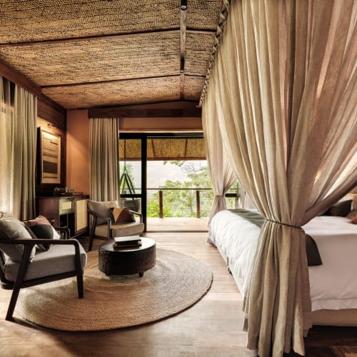 Luxuriöses Schlafzimmer in einer Lodge mit Himmelbett, umgeben von Vorhängen, mit Sitzbereich und Balkon mit Blick auf grüne Natur.