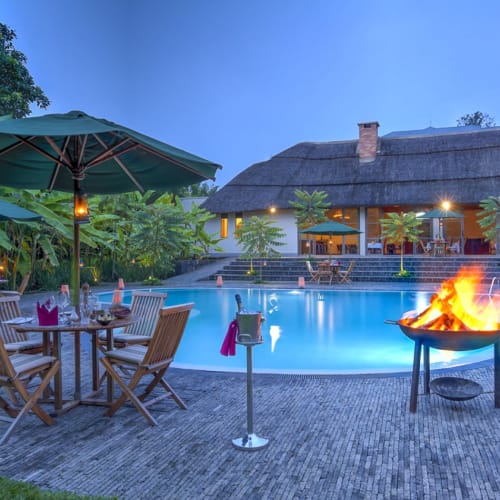 Außergewöhnliches Abendessen am Pool bei Dämmerung mit Holztischen und Stühlen unter grünen Sonnenschirmen, einem brennenden Feuerkorb und einem Strohdachgebäude im Hintergrund, umgeben von üppigem Grün.