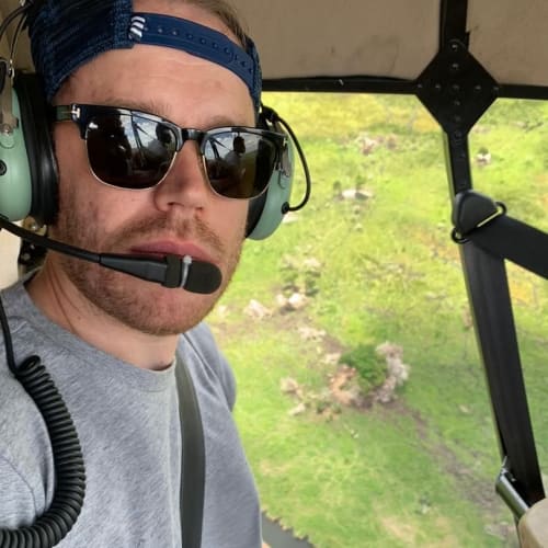 Mann mit Sonnenbrille, blauer Kappe und Headset mit Mikrofon sitzt in einem Hubschrauber und fliegt über eine grüne Landschaft mit Steinflecken.