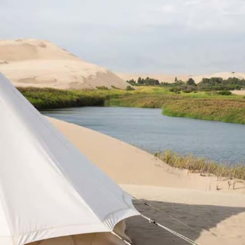 Ein weißes Zelt in der Nähe eines Flusses mit Sanddünen im Hintergrund unter einem bewölkten Himmel.