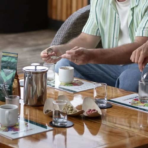 Drei Personen sitzen um einen Holztisch und bereiten Kaffee mit einer French Press und verschiedenen Kaffeezubehör vor in einem gemütlichen Innenraum.