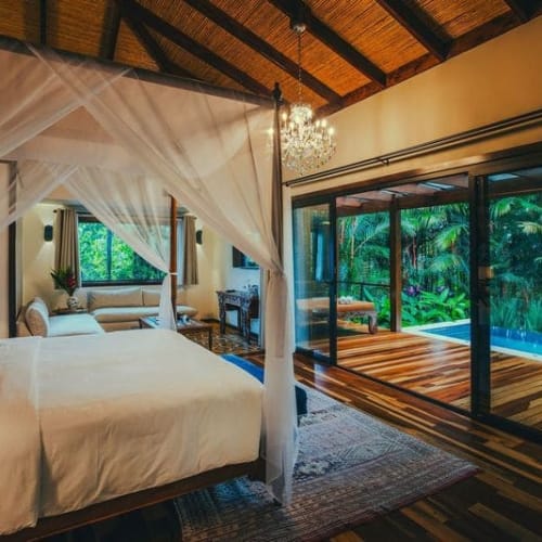 Luxuriöses Schlafzimmer mit Himmelbett, weißen durchsichtigen Vorhängen, einer großen Glastür, die zu einer Holzterrasse und üppigem tropischem Grün führt.