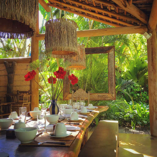 Outdoor dining area with a wooden table set for breakfast, surrounded by lush green tropical plants and rustic wooden beams, featuring a vase with red flowers and wicker pendant lights.