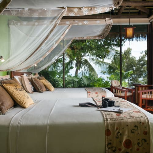 Luxurious bedroom with a canopy bed, decorated pillows, and an ocean view in a tropical lodge.
