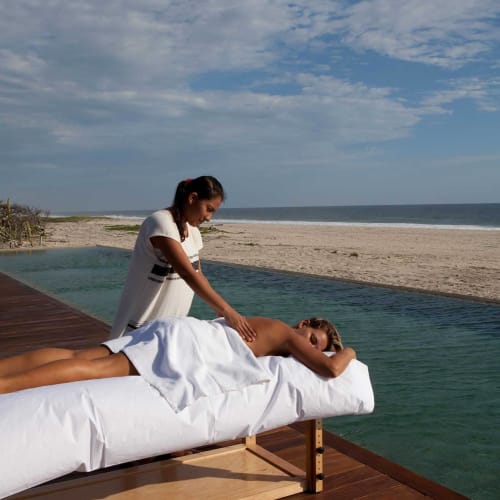 Eine Person erhält eine Massage auf einem weißen Massagetisch am Strand mit Meer und teilweise bewölktem Himmel im Hintergrund.