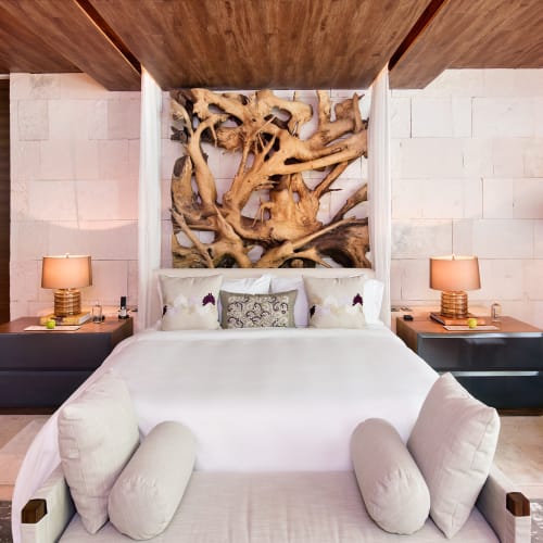 Modernes Hotelzimmer mit großem Bett, Holzdecken, zwei Nachttischen mit Lampen, einem dekorativen Holzkunstwerk über dem Kopfteil und einem Sitzbereich mit Kissen am Fußende des Bettes.