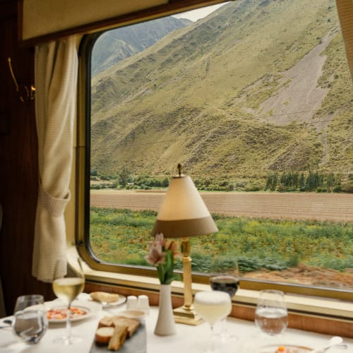 Ein Esstisch in einem Zug mit Tellern mit Essen, Weingläsern, einer kleinen Blumenvase und einer Lampe vor einem großen Fenster mit Blick auf grüne Felder und bergige Landschaft.