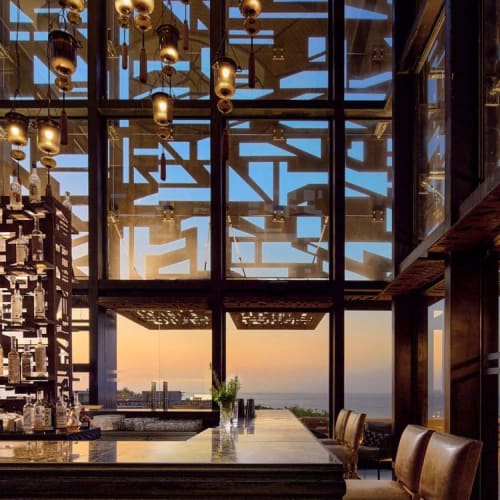 Innenraum einer Bar mit einer großen Holztheke, zwei hohen Lederstühlen, hängenden Laternenlampen und großen Fenstern mit Blick auf den Sonnenuntergang über dem Ozean.