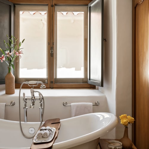 A cozy bathroom with a freestanding bathtub placed in front of a window with wooden frames and open black shutters, decorated with a vase of pink lilies on the windowsill and a side table with a yellow flower vase.
