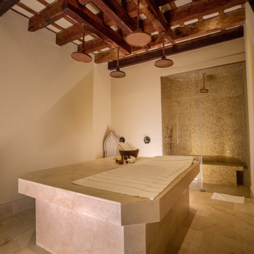 Innenansicht eines Spa-Zimmers mit einem großen Massagetisch aus Stein, Holzdeckenbalken, vier runden, an der Decke montierten Duschköpfen und einem gefliesten Duschbereich in der Ecke.