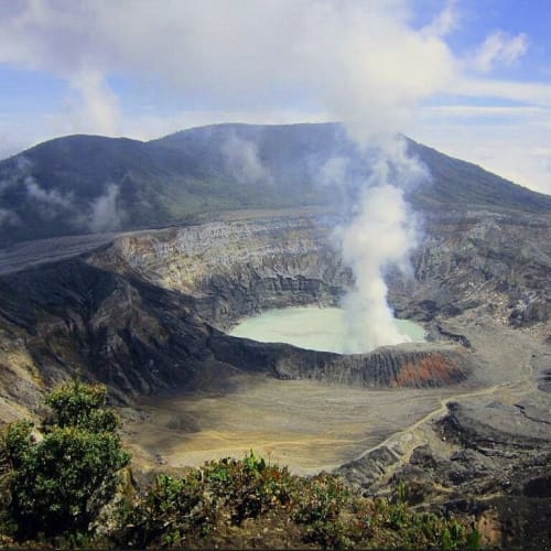 A smoking volcanic crater surrounded by rocky terrain and green vegetation under a partly cloudy sky.