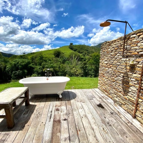 Outdoor bathtub and wooden bench on a wooden deck next to a stone wall with an overhead shower, surrounded by green hills under a partly cloudy blue sky.