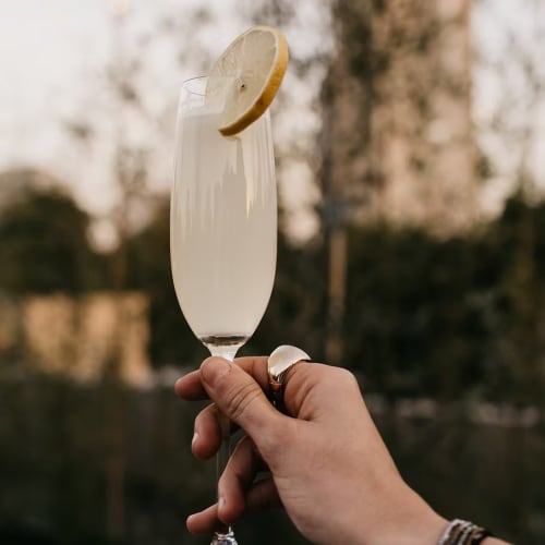 Hand hält ein Champagnerglas mit einem klaren, hellen Getränk und einer Zitronenscheibe als Garnitur im Freien vor unscharfem Baumhintergrund.