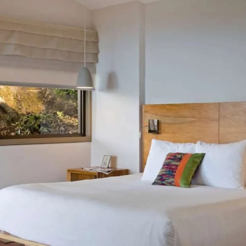 Minimalist bedroom featuring a wooden headboard queen-size bed with white linens and a colorful accent pillow next to a small wooden nightstand under a window with a beige roman shade.