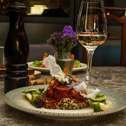 Ein schön angerichteter Teller mit einem Glas Weißwein, einem Pfefferstreuer und einem kleinen Blumenstrauß aus lila Blumen in einem Restaurant.