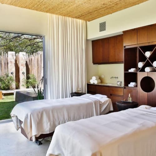Ein Spa-Massageraum mit zwei Massageliegen, die mit weißen Laken bedeckt sind, Holzmöbeln und Glastüren, die zu einem Außenbereich mit Liegestühlen und Grünflächen führen.