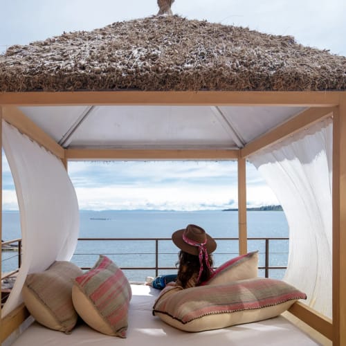 Eine Person sitzt unter einem Strohdach-Pavillon mit Blick auf ein ruhiges Meer, umgeben von großen Kissen und weißen transparenten Vorhängen.