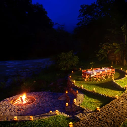 Außenbereich mit Essbereich bei Pacuare Lodge am Abend, beleuchtet von Kerzen und einem Lagerfeuer, in der Nähe eines Flusses und umgeben von üppigen Bäumen.