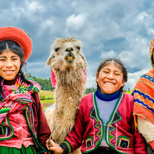 Three smiling children in colorful traditional Peruvian clothing with a llama under a cloudy sky.