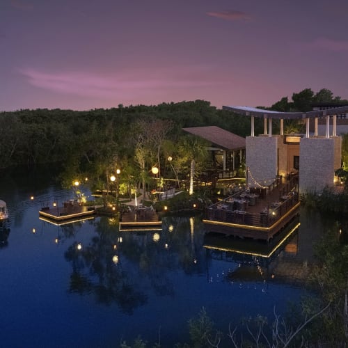 Eine ruhige Abendszene im Banyan Tree Mayakoba mit beleuchteten schwimmenden Plattformen und einem Boot auf ruhigem Wasser, umgeben von dichtem Grün unter einem violetten Abendhimmel.