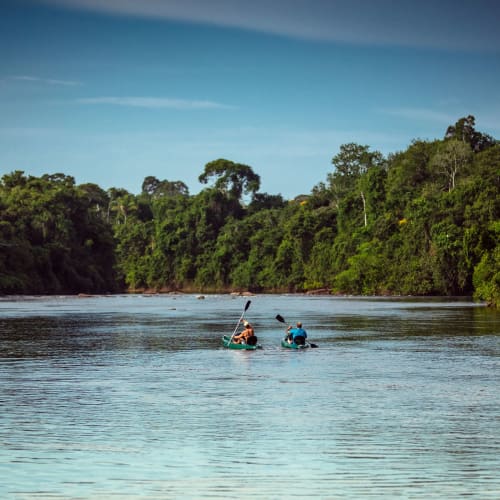 Zwei Personen beim Kanufahren auf einem breiten Fluss, umgeben von dichtem grünem Wald unter klarem blauem Himmel.