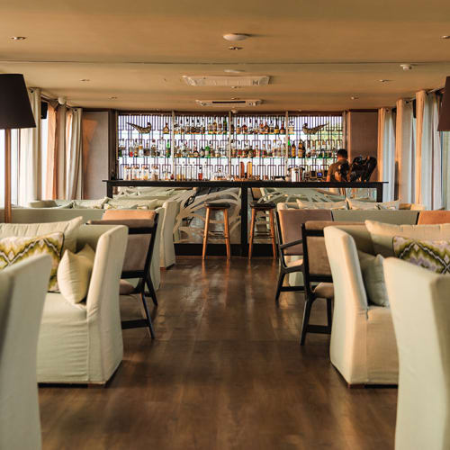 Eleganter Lounge-Bereich mit weißen Sessel und Sofas, symmetrisch angeordnet, im Hintergrund eine Bar mit Flaschen und große Fenster mit Vorhängen.