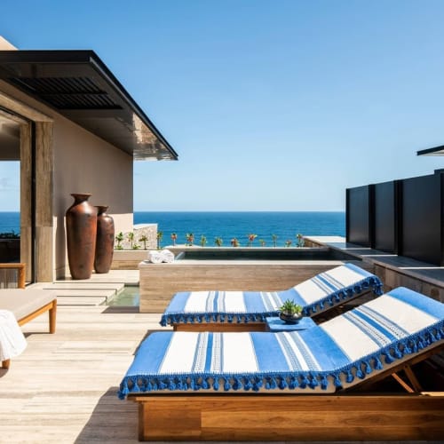 Moderne Außenterrasse mit zwei Holz-Liegestühlen mit blau-weißen gestreiften Polstern, einem Holzsofa mit Kissen, großen Vasen und einem Infinity-Pool mit Blick auf den Ozean unter klarem blauem Himmel.