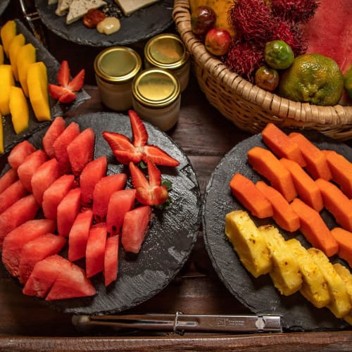 Freshly cut watermelon, cantaloupe, papaya, and pineapple arranged on black plates with jars and tropical fruits in the background.