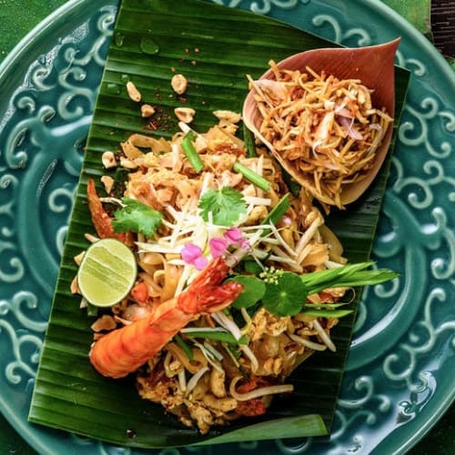 Ein Teller Pad Thai-Nudeln, garniert mit Garnelen, Limettenscheibe, frischen Kräutern und Sojasprossen, serviert auf einem Bananenblatt mit dekorativen Blütenblättern und Lotusknospen an der Seite.