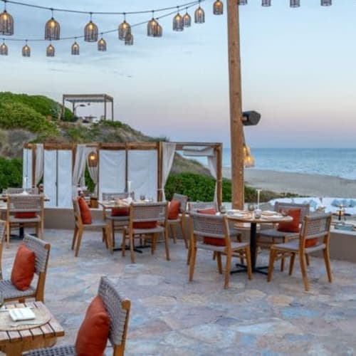 Außenbereich eines Restaurants am Meer mit Tischen und Stühlen auf einem Steinboden, mit Blick auf den Strand und das Meer unter Lichterketten bei Dämmerung.