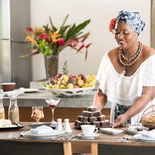 Eine Frau in traditioneller Kleidung serviert Desserts an einem Tisch mit Früchten und Blumen im Hintergrund.