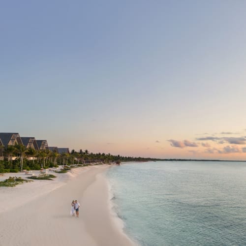 Ein Paar spaziert bei Sonnenuntergang an einem breiten Sandstrand entlang, neben einer Reihe von Villen des Resorts mit Palmen.