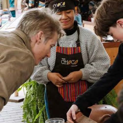 Eine Frau mit einem CHOC-Schurz und Stirnband zeigt zwei Männern beim Schokoladengenuss an einem Marktstand.
