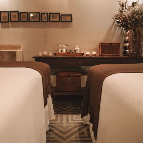 Zwei Massageliegen mit braunen Decken in einem Spa-Raum mit einem Regal, das Kerzen, Dekorationsgegenstände und getrocknete Blumen in einer Vase vor einer beigen Wand enthält.