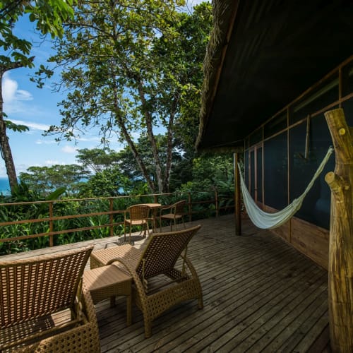 Eine geräumige Holzterrasse mit Rattanstühlen, einer Hängematte und Blick auf üppige grüne Bäume und den Ozean im Hintergrund.