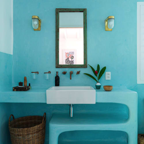 Bathroom with turquoise walls, a white rectangular sink with wall-mounted bronze faucets, a rustic frame mirror, brass wall sconces, and woven baskets underneath a built-in shelf.