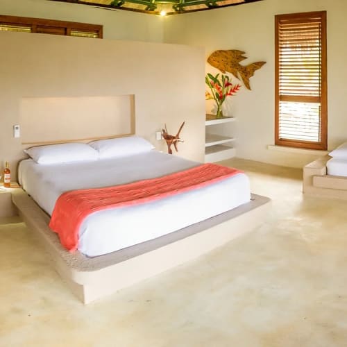 Spacious minimalist bedroom with two beds, one large bed with white bedding and a red throw, and a smaller bed with white and green bedding, set against beige walls and concrete flooring with wooden window shutters.