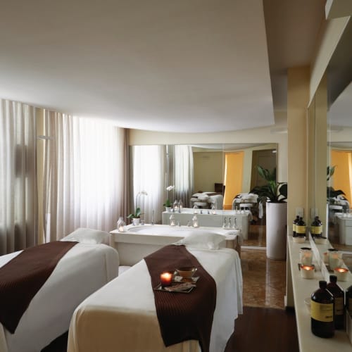 Ein luxuriöser Hotel-Spa-Raum mit zwei Massagebetten, Kerzen und einem großen Spiegel, der den Raum reflektiert, in einem hellen Raum mit großen Fenstern und Vorhängen.