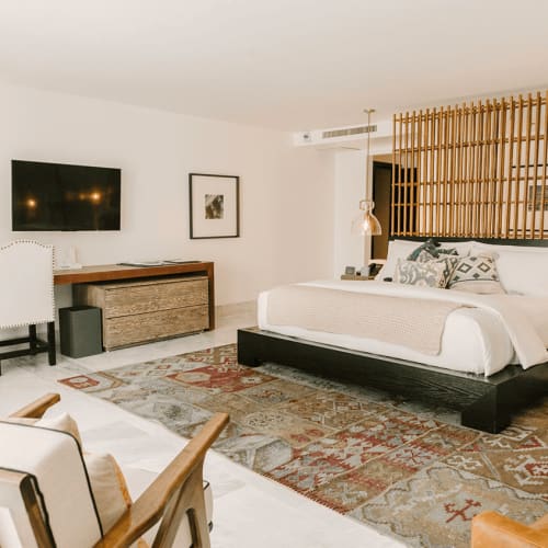 Geräumiges Luxusschlafzimmer mit großem Bett und dekorativen Kissen, einem hölzernen Lattenkopfteil, einem gemusterten Teppich, Schreibtisch mit Stuhl und moderner Beleuchtung.