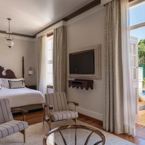Elegantes Schlafzimmer mit großem Bett, antiken Stühlen und Blick auf einen Außenpool durch offene Glastüren.