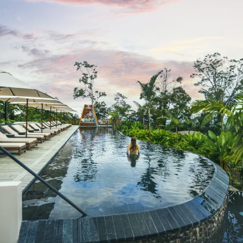 Person schwimmt in einem Infinity-Pool, umgeben von Liegestühlen mit Sonnenschirmen auf der einen Seite und üppigem Grün auf der anderen Seite unter einem pastellfarbenen Himmel bei Sonnenuntergang oder Sonnenaufgang.