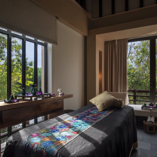 Ein ruhiger Spa-Behandlungsraum mit einer Massageliege, die mit einem schwarzen Tuch und einem bunten Läufer bedeckt ist, Tageslicht, das durch große Fenster auf grüne Bäume draußen fällt.