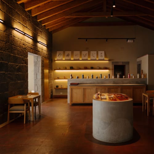 A cozy, dimly lit restaurant interior with wooden furniture, brick and stone walls, and shelves with various items behind the counter.