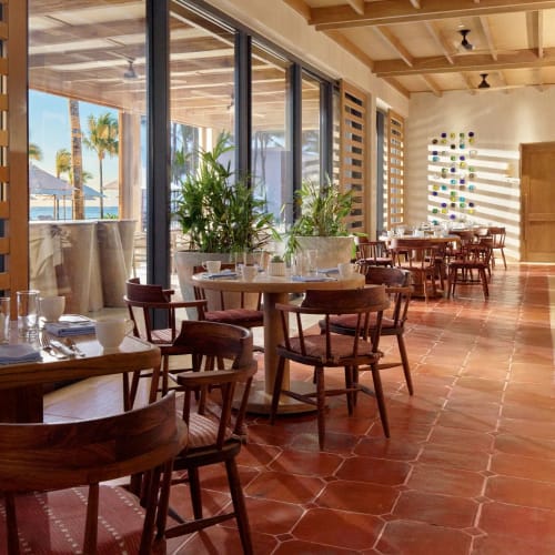 Heller Innenbereich eines Restaurants mit Holztischen und Stühlen, großen Glasfenstern mit Blick auf eine Außenterrasse mit Palmen und buntem Fliesenboden im Morgenlicht.