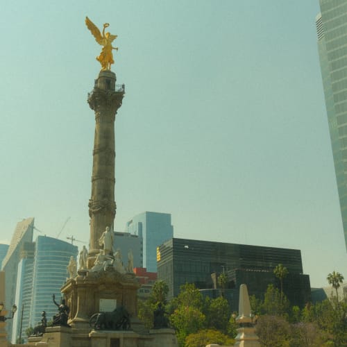 Das Unabhängigkeitsdenkmal in Mexiko-Stadt mit einer goldenen Engelsfigur auf einer hohen Säule, umgeben von modernen Wolkenkratzern und Grünflächen.